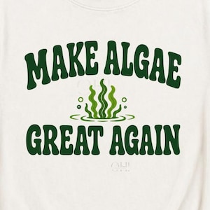 Könnte beinhalten: Ein cremefarbenes Kurzarm-T-Shirt mit dunkelgrünem Text "MAKE ALGAE GREAT AGAIN" und einer Grafik aus grünem Seetang in der Mitte. Das Shirt hat einen Rundhalsausschnitt und eine Standardpassform, dargestellt als flach liegendes Kleidungsstück.