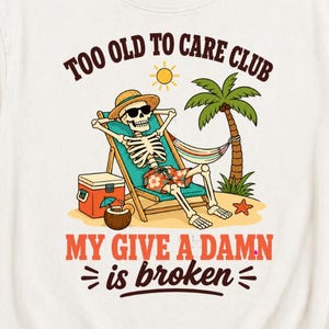 May include: An off-white t-shirt featuring a graphic of a skeleton wearing sunglasses and a straw hat, relaxing in a beach chair under a palm tree. The text reads, "TOO OLD TO CARE CLUB" and "MY GIVE A DAMN is broken" in brown and red lettering.