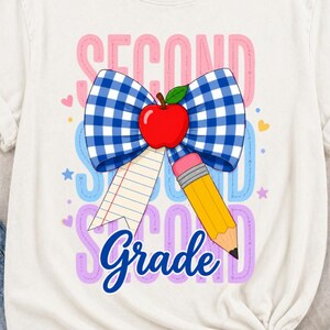 Può includere: Una t-shirt a maniche corte color crema con una grafica raffigurante un fiocco a quadretti blu e bianco, una mela rossa e una matita gialla. Il testo "SECOND Grade" è stampato in blu, circondato da piccoli cuori e stelle.