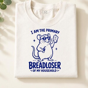 May include: Cream-coloured crew neck t-shirt featuring a navy blue cartoon rat wearing sunglasses, holding a drink and bread. The text reads "I AM THE PRIMARY BREADLOSER OF MY HOUSEHOLD."