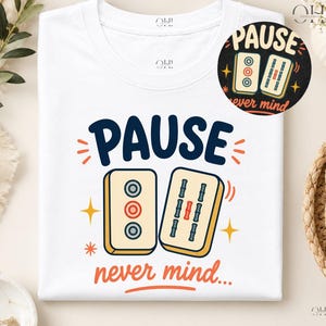 Könnte beinhalten: Ein weißes T-Shirt mit einem Aufdruck, der den dunkelblauen Schriftzug "PAUSE" über zwei Mahjong-Steinen zeigt. Darunter steht in orangefarbenen Buchstaben "never mind..." mit kleinen Sternen. Das Design enthält einen runden Ausschnitt.
