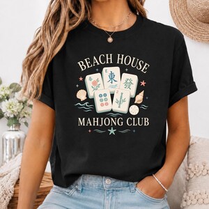 May include: Black t-shirt with the words "BEACH HOUSE MAHJONG CLUB" in white. The design includes Mahjong tiles, seashells, and stars. The shirt is a casual style.