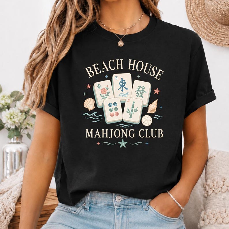 May include: Black t-shirt with the words "BEACH HOUSE MAHJONG CLUB" in white. The design includes Mahjong tiles, seashells, and stars. The shirt is a casual style.