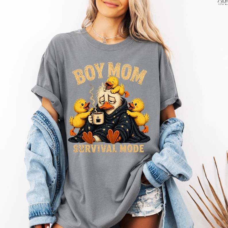 May include: Gray t-shirt with the text "BOY MOM" in gold letters above a cartoon duck wearing a blanket and holding a coffee cup. Three yellow ducklings surround the duck. The text "SURVIVAL MODE" is below the duck.