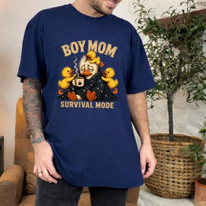 May include: Navy blue t-shirt with the text "BOY MOM" in gold lettering above a cartoon duck wearing a blanket and holding a coffee cup, surrounded by yellow ducklings. The phrase "SURVIVAL MODE" is printed below the duck.