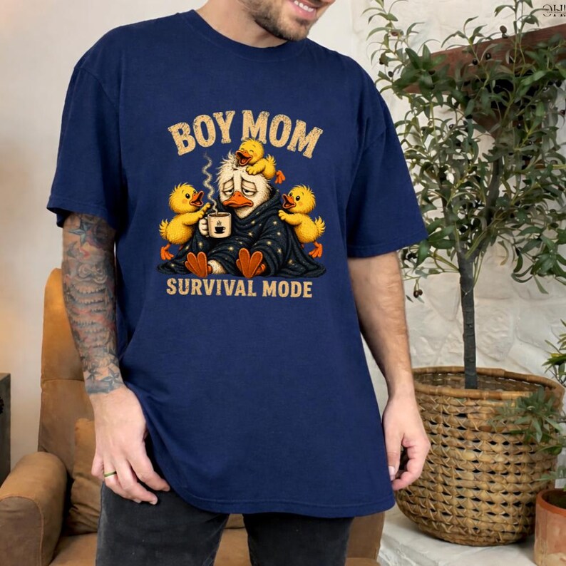 May include: Navy blue t-shirt with the text "BOY MOM" in gold lettering above a cartoon duck wearing a blanket and holding a coffee cup, surrounded by yellow ducklings. The phrase "SURVIVAL MODE" is printed below the duck.