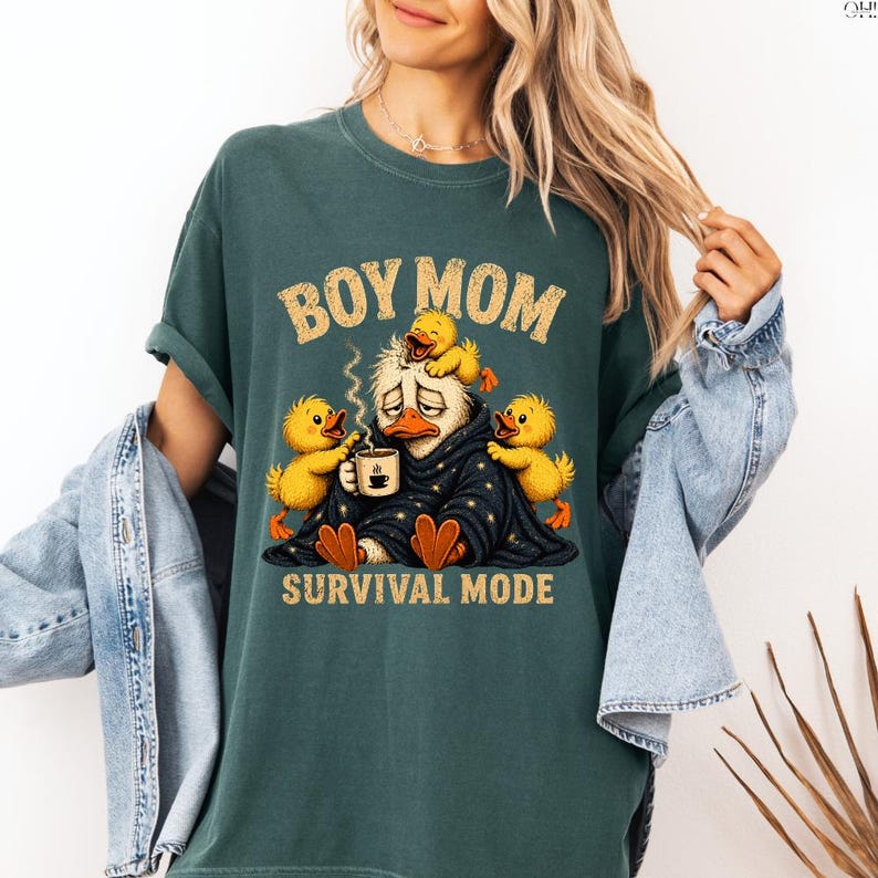 May include: Dark teal t-shirt with the text "BOY MOM" in a distressed font. The graphic features a tired duck character wrapped in a blanket, holding a coffee cup, surrounded by yellow ducklings. The words "SURVIVAL MODE" are below the graphic.