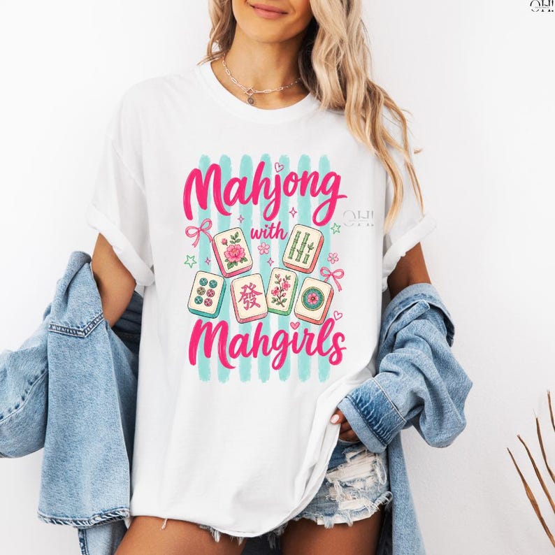 May include: White t-shirt with a colorful graphic design. The design features the words "Mahjong with Mahgirls" in pink script, along with illustrations of Mahjong tiles. The background has vertical blue stripes.