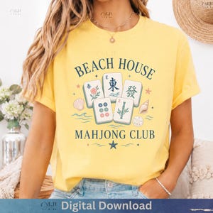 May include: Yellow t-shirt with a graphic design featuring the words "BEACH HOUSE MAHONG CLUB" and illustrations of Mahjong tiles, seashells, and stars. The shirt has short sleeves and a crew neck.