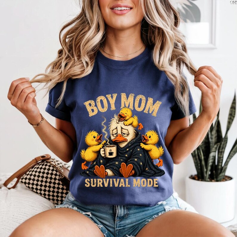 May include: Navy blue t-shirt with the text "BOY MOM" in gold letters. The graphic depicts a tired duck surrounded by yellow ducklings, wrapped in a blanket, holding a coffee cup. The words "SURVIVAL MODE" are below the graphic.