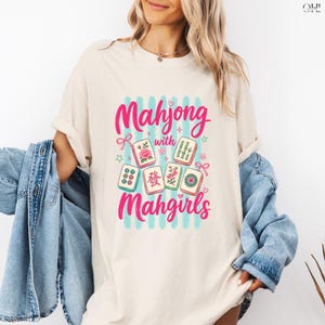May include: Cream-colored t-shirt featuring a graphic design with the words "Mahjong with Mahgirls" in pink script. The design includes illustrations of Mahjong tiles with floral and bamboo motifs, set against a backdrop of blue stripes.