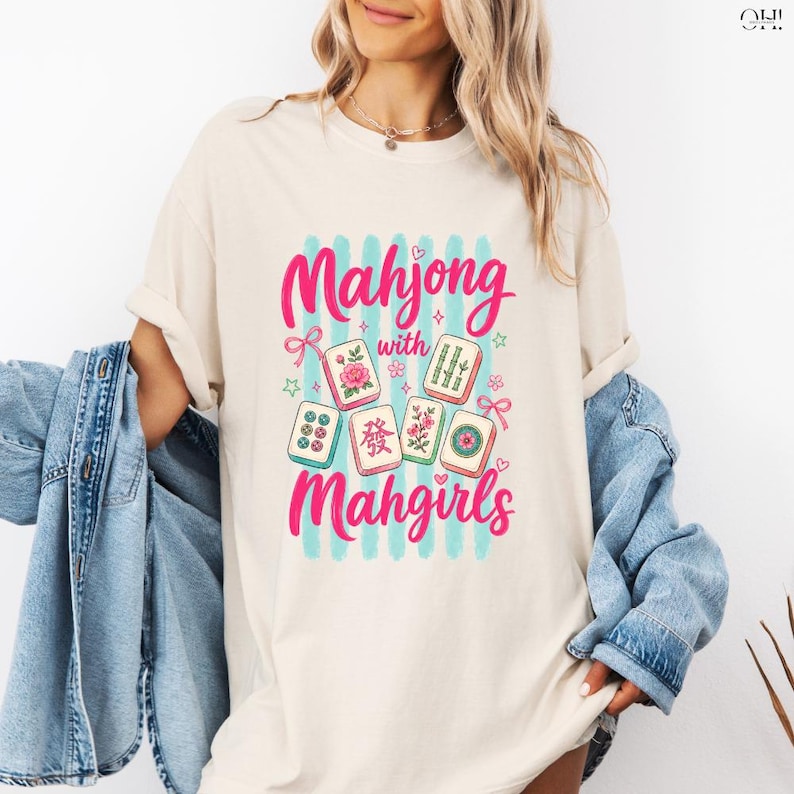 May include: Cream-colored t-shirt featuring a graphic design with the words "Mahjong with Mahgirls" in pink script. The design includes illustrations of Mahjong tiles with floral and bamboo motifs, set against a backdrop of blue stripes.
