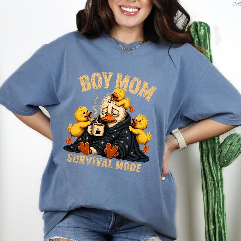 May include: A blue t-shirt with the words "BOY MOM" and "SURVIVAL MODE" in a distressed font. The graphic features a tired duck wrapped in a blanket, holding a coffee cup, surrounded by three yellow ducklings.