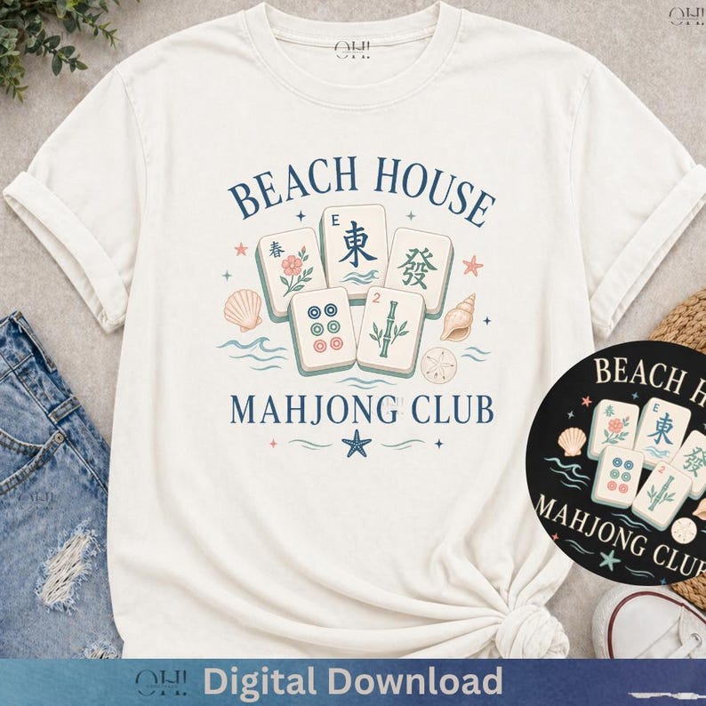 May include: Cream-colored t-shirt featuring a "Beach House Mahjong Club" design. The design includes Mahjong tiles, seashells, and the text "Beach House Mahjong Club" in navy blue. The shirt is styled with rolled-up sleeves and denim jeans.