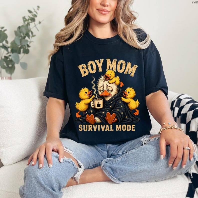 May include: Black t-shirt with the text "BOY MOM" above a cartoon duck wearing a blanket and holding a coffee cup, surrounded by yellow ducklings. The phrase "SURVIVAL MODE" is printed below the graphic.
