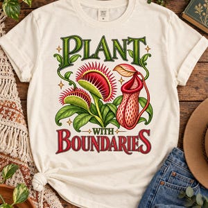 May include: A cream-coloured short-sleeve t-shirt featuring a graphic of two green Venus flytraps with red interiors and a red-patterned pitcher plant. The text reads "PLANT" in green at the top and "WITH BOUNDARIES" in red at the bottom.