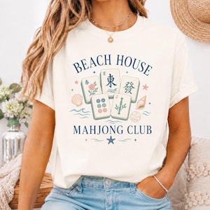 May include: Cream-colored t-shirt with a graphic design featuring the text "BEACH HOUSE MAHONG CLUB". The design includes illustrations of Mahjong tiles, seashells, and floral elements in shades of blue, pink, and green.