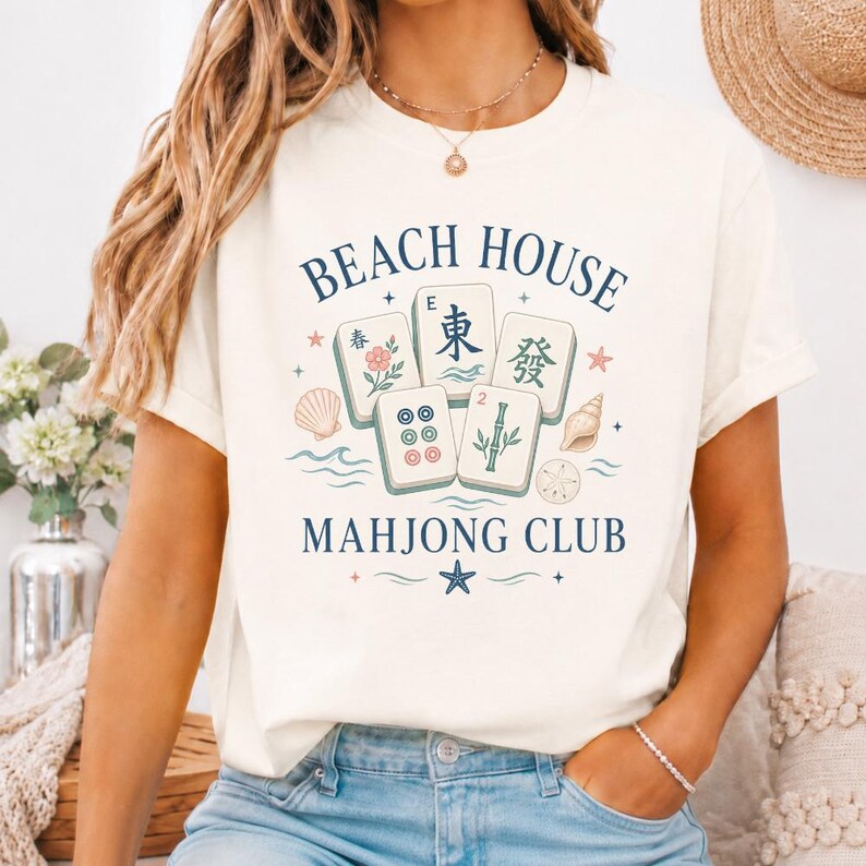 May include: Cream-colored t-shirt with a graphic design featuring the text "BEACH HOUSE MAHONG CLUB". The design includes illustrations of Mahjong tiles, seashells, and floral elements in shades of blue, pink, and green.