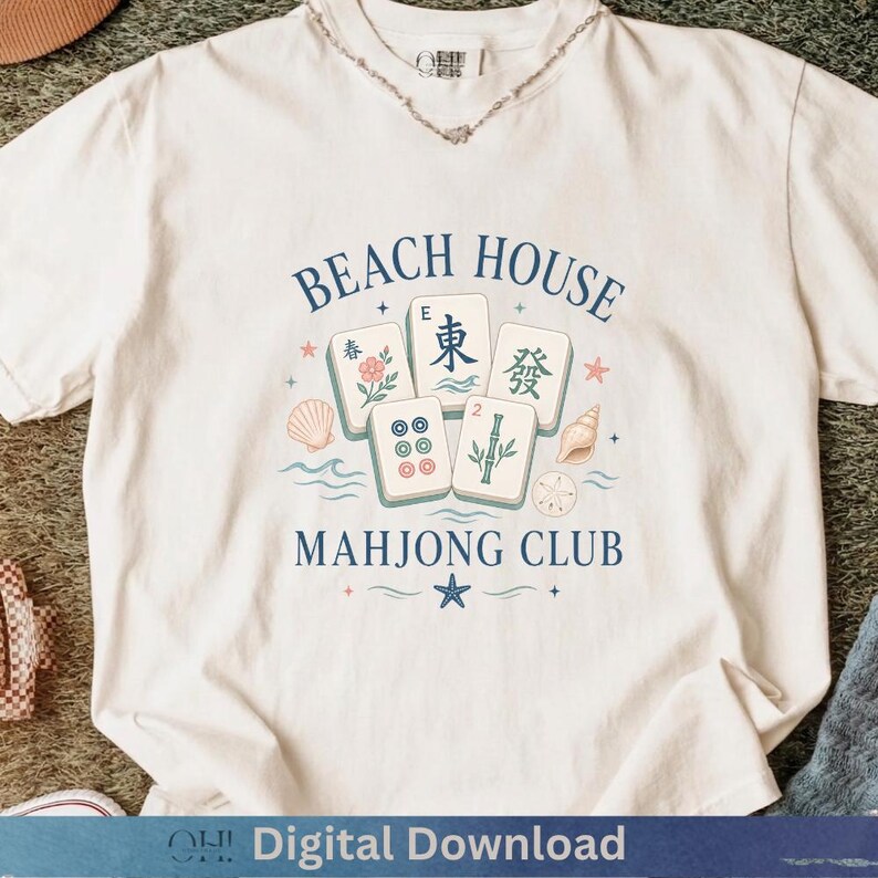 May include: Cream-colored t-shirt with a graphic design featuring the words "BEACH HOUSE MAHONG CLUB" and illustrations of Mahjong tiles, seashells, and a star. The design is in shades of blue, green, and pink.