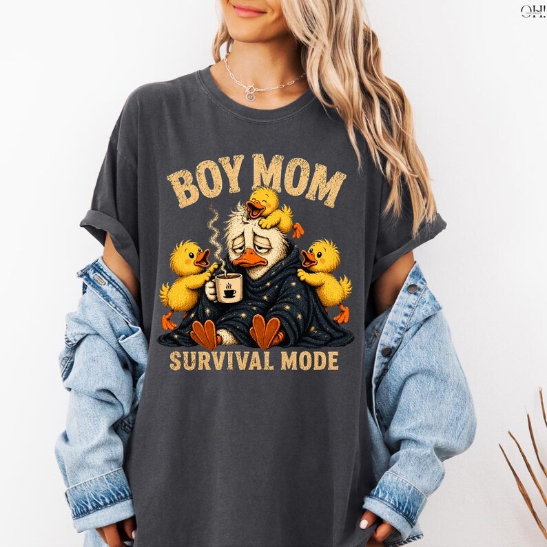 May include: Dark gray t-shirt with the text "BOY MOM" above an illustration of a duck wrapped in a blanket, surrounded by three yellow ducklings. The text "SURVIVAL MODE" is below the illustration.