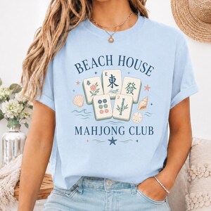 May include: Light blue t-shirt with the text "BEACH HOUSE MAHONG CLUB". The shirt features a graphic of Mahjong tiles, seashells, and the Chinese characters for East and Spring.