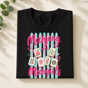May include: Black t-shirt featuring the text "Mahjong with Mahgirls" in pink script, with light blue vertical stripes and illustrated Mahjong tiles. The tiles have floral and bamboo designs.