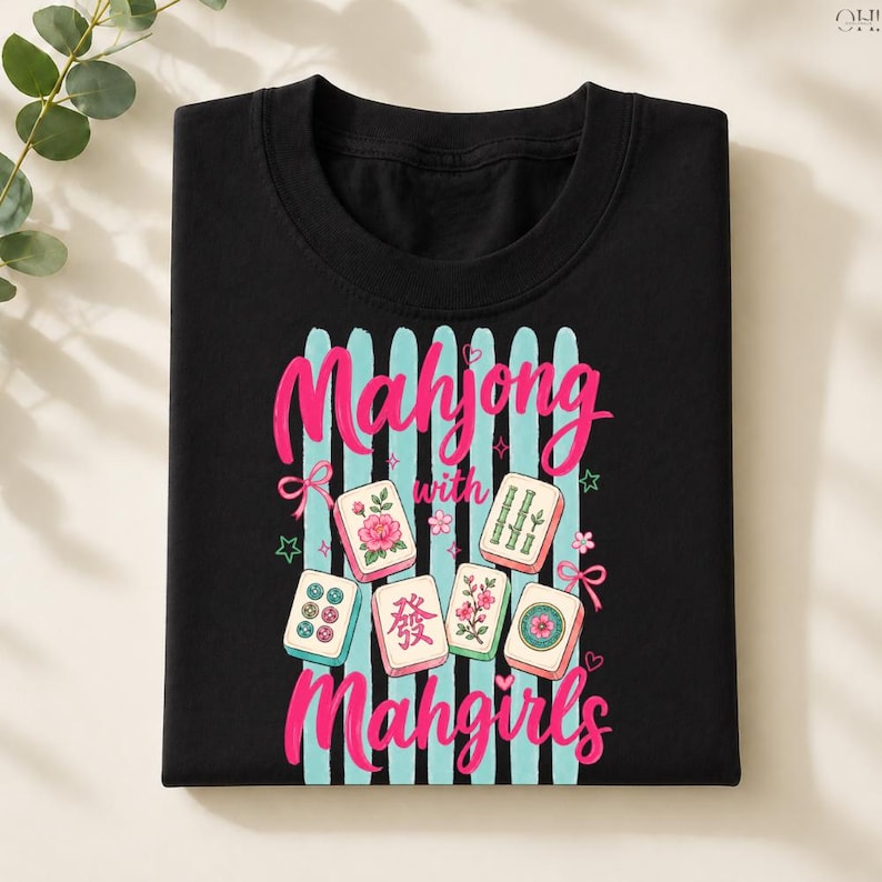 May include: Black t-shirt featuring the text "Mahjong with Mahgirls" in pink script, with light blue vertical stripes and illustrated Mahjong tiles. The tiles have floral and bamboo designs.