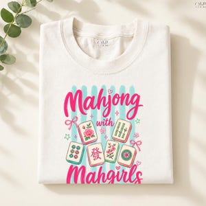 May include: White crew neck t-shirt featuring the words "Mahjong with Mahgirls" in pink script. The design includes illustrations of colorful Mahjong tiles with floral and bamboo motifs, set against a backdrop of blue vertical stripes.