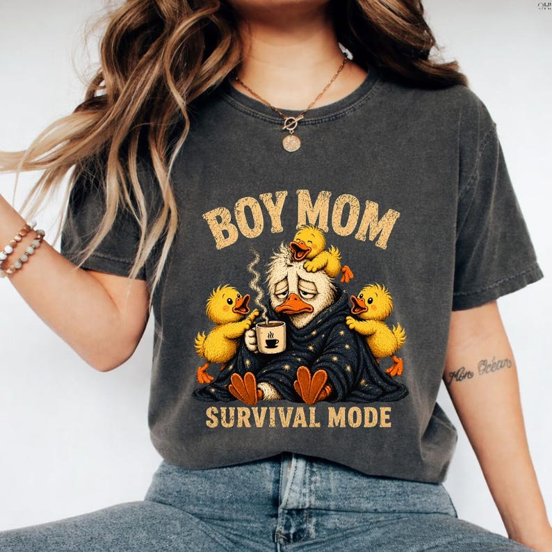 May include: Dark gray t-shirt with the text "BOY MOM" above a cartoon duck character wrapped in a blanket, holding a coffee cup, surrounded by yellow ducklings. The text "SURVIVAL MODE" is below the duck.