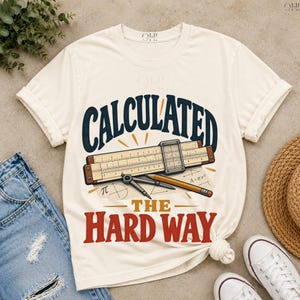 Puede incluir: Una camiseta de color crema con un gráfico de una regla de cálculo, un compás y un lápiz. El texto "CALCULATED THE HARD WAY" aparece en azul oscuro y rojo, junto a símbolos matemáticos que incluyen el signo pi y la fórmula A=πr².