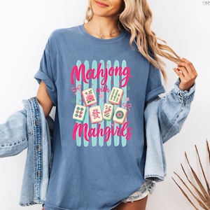May include: A blue t-shirt with the text "Mahjong with Mahgirls" in pink and white. The shirt features a graphic of Mahjong tiles with floral and bamboo designs. The shirt is a casual style.