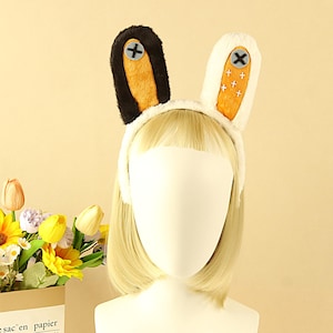 May include: A white headband with plush bunny ears. One ear is white with orange accents and black cross-stitch details, the other is black with orange accents and a black cross-stitch. The headband is on a mannequin head with blonde hair.