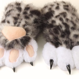 Peut inclure: Une paire de gants en forme de pattes d'animaux en peluche. Les gants présentent un motif de fourrure grise et noire tachetée sur le dessus, avec de la fourrure blanche en dessous. Les coussinets sont de couleur beige clair et les griffes sont noires.