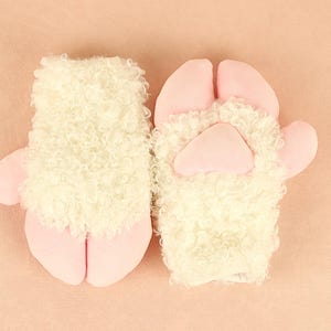 Peut inclure: Une paire de mitaines fantaisie en forme de mouton. Les mitaines sont blanches avec une texture duveteuse, ressemblant à de la laine, et présentent des détails roses en forme de pattes. Le fond est de couleur pêche clair.