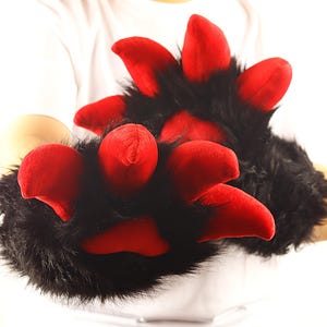 May include: Black and red plush paw gloves. The gloves have black fur with bright red padded claws. The gloves are designed to look like animal paws. The gloves are being held up by a person wearing a white shirt.