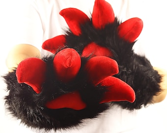 May include: Black and red plush paw gloves. The gloves have black fur with bright red padded claws. The gloves are designed to look like animal paws. The gloves are being held up by a person wearing a white shirt.