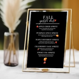 May include: A black menu sign titled FALL SPRITZ BAR in a thin gold metal frame. The menu lists five cocktails: Apple Cider Spritz, Pumpkin Spice Spritz, Maple Bourbon Spritz, Cranberry Sage Spritz, and Smoky Fig Spritz with small icons.
