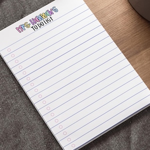 May include: A white notepad with blue lined pages and pink circles for checkboxes. The notepad has the text "MRS. HANNAH'S TO DO LIST!" in colorful lettering at the top. The notepad is on a wooden desk next to a laptop and a pen.
