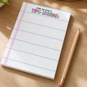 May include: A rectangular notepad with white paper featuring horizontal light blue lines and a vertical pink margin line. The top of the page displays the text "From the desk of... Mrs. Hannah" in colorful, rounded lettering. A wooden pencil lies nearby.