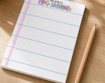 May include: A rectangular notepad with white paper featuring horizontal light blue lines and a vertical pink margin line. The top of the page displays the text "From the desk of... Mrs. Hannah" in colorful, rounded lettering. A wooden pencil lies nearby.