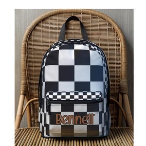 May include: Black and white checkered backpack with the name "Bennett" in brown lettering. The backpack has a front pocket with a zipper and a black handle. The bag is sitting on a wicker chair.