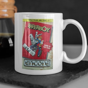 Könnte beinhalten: Weiße Keramiktasse mit einem roten und weißen Bild einer Videospielkassette für Paperboy auf dem Spectrum 48k/128k +2. Das Bild zeigt einen Jungen, der Fahrrad fährt und Zeitungen ausliefert. Der Text "Game of the Year" befindet sich auf der Kassette. Der Text "Encore" befindet sich am unteren Rand des Bildes.