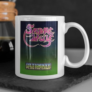 Könnte beinhalten: Weiße Keramiktasse mit einer bunten Grafik des Textes "Sabre Wulf" und den Worten "Ultimate Play the Game".