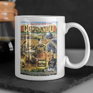 Könnte beinhalten: Weiße Keramiktasse mit einem bunten Bild eines Videospielcharakters, der eine Waffe hält. Der Text "The smash hit No. 1 arcade game! Commando" ist auf die Tasse gedruckt.