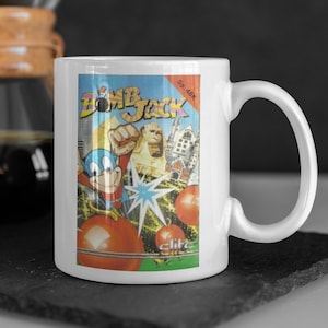 Könnte beinhalten: Weiße Keramiktasse mit einem bunten Bild eines Videospielcharakters, einem blau-roten Superhelden, der Bomben wirft. Der Text "Bomb Jack" und "Sp. 48K" ist auf der Tasse sichtbar.