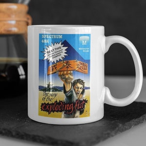 Puede incluir: Taza de cerámica blanca con una imagen colorida de un karateca rompiendo una tabla de madera. La imagen es del videojuego "The Way of the Exploding Fist" para el Spectrum 48K.