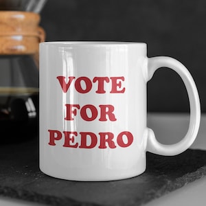 May include: White ceramic mug with the text "VOTE FOR PEDRO" in red, with a coffee maker in the background. The mug is on a black surface.