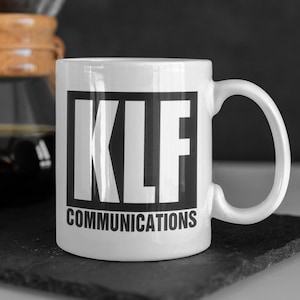 Può includere: Tazza in ceramica bianca con un quadrato nero che contiene il testo "KLF COMMUNICATIONS" in lettere bianche.