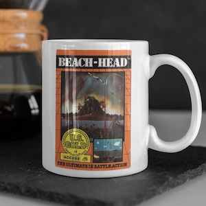 Könnte beinhalten: Weiße Keramik-Kaffeetasse mit einer Grafik des Videospiels Beach-Head, auf einem roten und orangenen Hintergrund mit einem gelben U.S. Gold-Logo. Der Text "The Ultimate in Battle Action" ist unter der Grafik gedruckt.