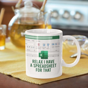 Puede incluir: Taza de cerámica blanca con un diseño de hoja de cálculo de Microsoft Excel verde y blanco. La taza tiene un asa negra y el texto "RELAX I HAVE A SPREADSHEET FOR THAT" impreso en la parte delantera.